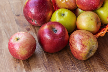 Red, green and yellow apples rolled out of a red wicker basket onto a brown wooden surface, side view from above