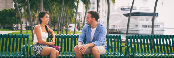 Fototapeta na wymiar Couple talking sitting on city bench urban lifestyle - young interracial people dating or going out on date meeting in summer Miami panoramic banner. Florida travel Asian woman, Caucasian man.