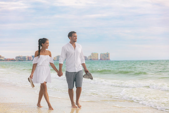 Beach couple romantic sunset walk Asian woman and Caucasian man relaxing walking on Florida vacation beach travel holidays wearing white dress and linen clothes. Happy interracial relationship.