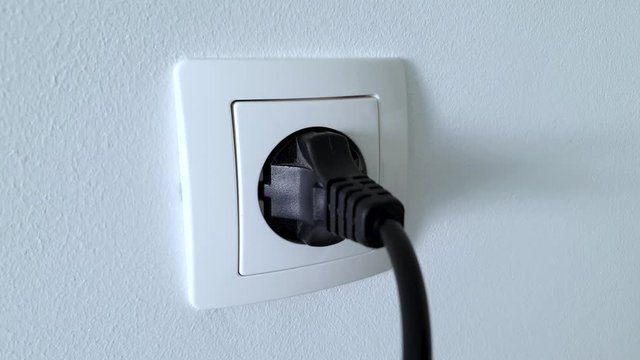 Connecting black electrical power plug with a hand inside a white plastic power socket on a white wall