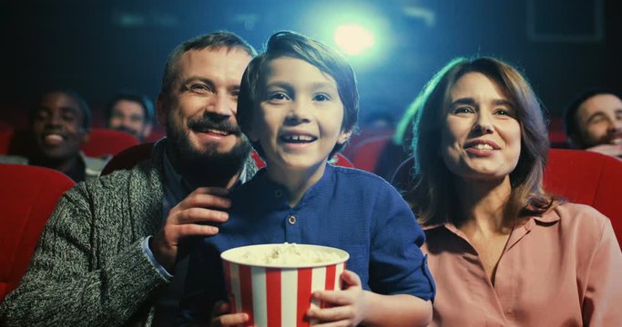 Portrait of teh happy small cute boy with popcorn in hands sitting in the cinema between cheerful parents and they laughing as watching comedy movie.
