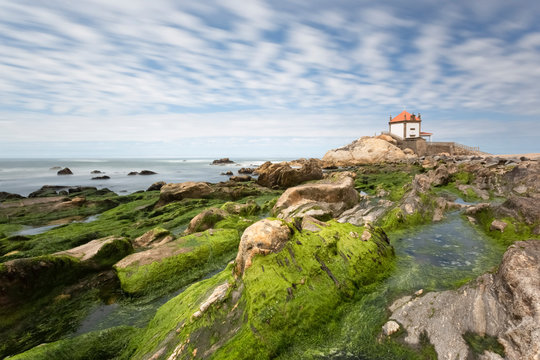 Chapel of Senhor da Pedra on rock formation by sea against cloudy sky