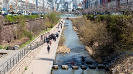 Timelapse people relax and wander along Seoul Cheonggyecheon stream recreation space with rocks and trees on banks against city highrise buildings zoom in