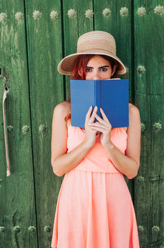 Portrait of redheaded young woman standing in front of green wooden, door covering mouth with a book