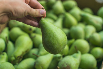 Human hand holding green pear in front of box full of pears after harvest.