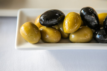 Variety Of Green And Black Marinated Olives Dish On Small Ceramic White Plate. Selective Focus. Close Up Of Tasty And Healthy Appetizer.