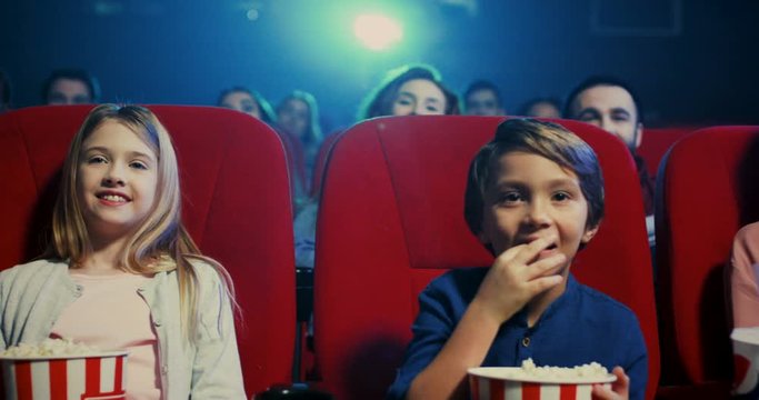 Cute small Caucasian children, boy and grl, eating popcorn and watching interesting film in the cinema with their parents.