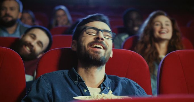 Close up of the handsome Caucasian young man in glasses watching a comedy movie in the cinema and eating popcorn while laughing loud.