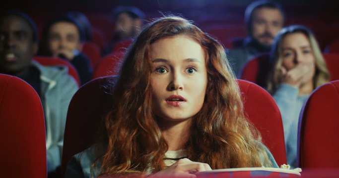 Close up of the Caucasian young attractive woman with red hair watching excited interesting action movie in the cinema and eating popcorn.