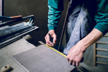 Close up on construction worker craftsman using tool for cutting ceramic tiles for laying installation to the bathroom making measurement marks using measuring tape and pen