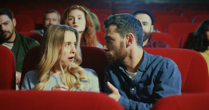 Caucasian young attractive boyfriend and girlfriend sitting together in the cinema and fighting because of the popcorn and people from behind rows asking them to be quietly.