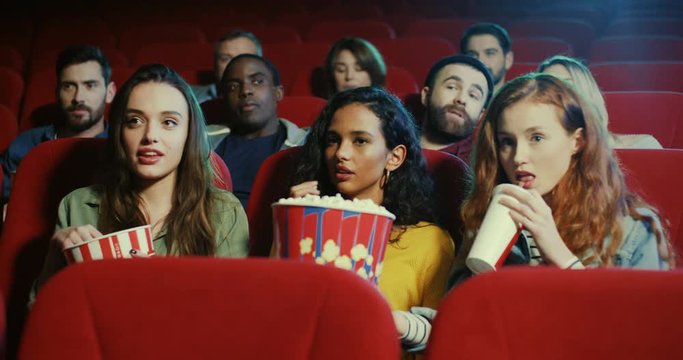 Portrait of the happy young Caucasian pretty girls friends watching a comedy movie in the cinema with popcorn, laughing and talking.