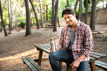 Obraz premium Model man holds a reusable aluminum bottle for water sitting in a park with trees.