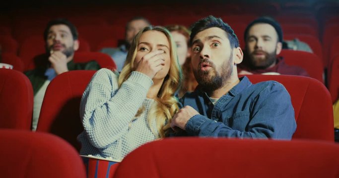 Caucasian young attractive boyfriend and girlfriend sitting in the cinema together and closing their eyes as being afraid becaude watching horror film.