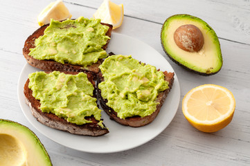 Healthy fat diet, nutritious snack and mexican food conceptual idea with smashed avocado into guacamole spread on rye bread toast surrounded by lemon and avocados isolated on white wood table