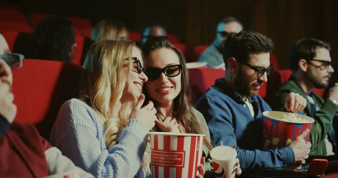 Young Caucasian beautiful and cheerful people in 3D glasses sitting in the cinema with popcorn and watching a movie while talking quietly.
