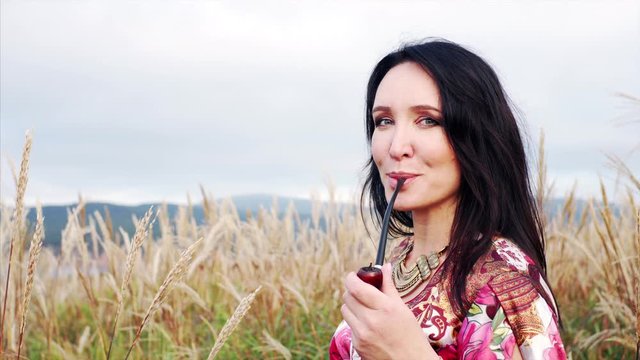 Portrait of attractive gipsy woman in colorful dress smoking pipe and looking into the camera with smile. Field with high grass. Summer