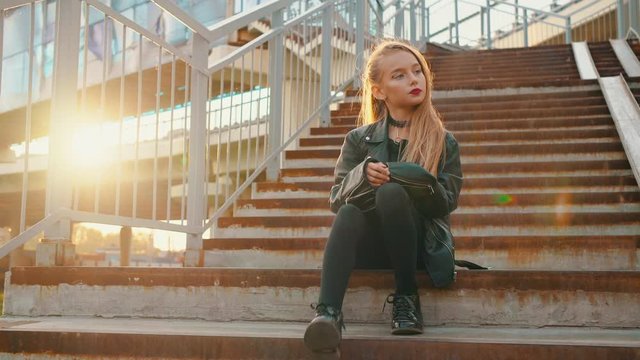 Lonely girl in black leather jacket sitting on stairs at sunshine background. Attractive girl in leather jacket on ground crossing stairs on golden sunflare background