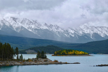Colourful island in Upper Kananaskis lake surrounded by snowy mountains on a foggy day. Autumn foliage of Canadian Rockies. Peter Lougheed Provincial Park. Alberta. Canada.