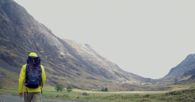 4K A Young Man in The Glen Coe Scottish Mountains. A Student Tourist in Yellow jacket and Backpack hiking on a Road within a Hilly Scenic destination in Rural Scotland.