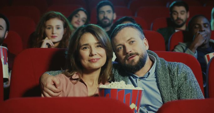Portrait shot of the Caucasian good looking married couple, man and woman sitting together in the cinema with popcorn, hugging and watching movie.