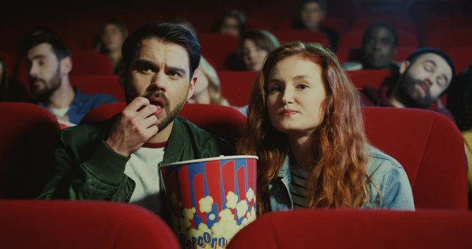 Handsome young Caucasian man asking something in the pretty red-haired woman, his girlfriend while they watching a film and eating a popcorn in the cinema.