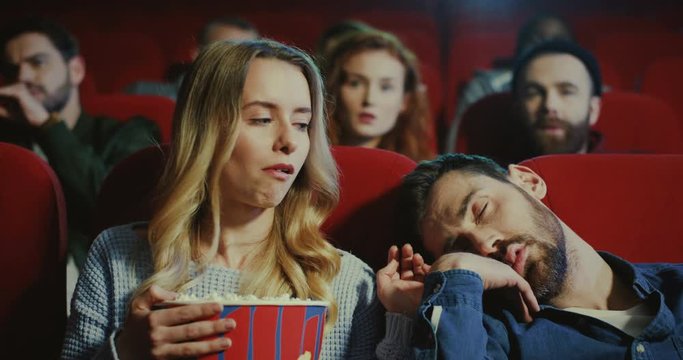 Portrait of the young pretty blond Caucasian woman watching a movie in the cinema with her boyfriend and he falling asleep, so she waking him up and telling to watch a film, but he is bored.