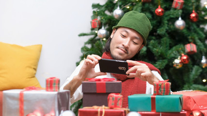 Obraz na płótnie Canvas Young asian man smiling while taking photo of Christmas present gift box, Happy asia blogger photo and recording vlog of Christmas gift and ornaments at celebration party