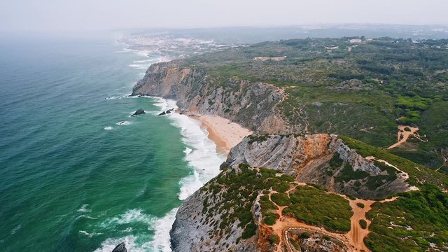 4K Aerial footage of Praia da Adraga beach and Atlantic coast. White ocean waves rolling towards the shore. Sintra, Portugal
