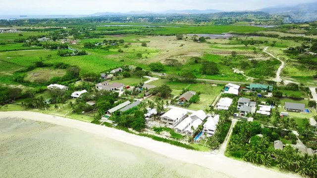 Aerial view: Sandy beachfront on pacific island with small houses, bures. Smoke from sugar cane fire and mountain in background