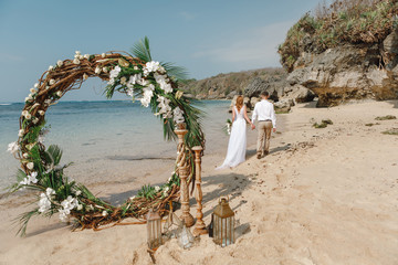 Happy groom and bride walking on whie sand beach  under the arch decorated with flowers in boho...