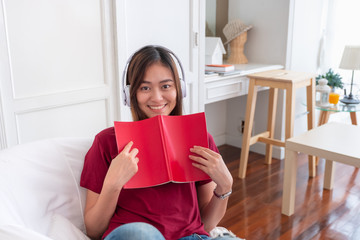 Asian woman listening music on headphone and reading book with relax pose at couch in living room at home.leisure lifestyle