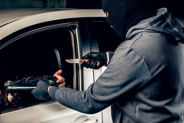 The robber in Balaclava threatening with a knife takes away a bag from the driver of a car. The...