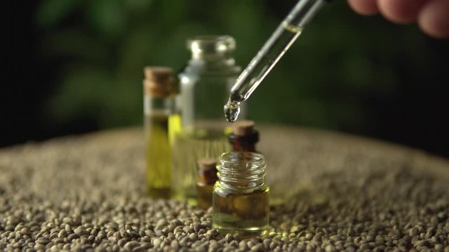 Macro close up of droplet pipette dosing biological and ecological oil of the hemp plant.  Pharmaceutical herbal cbd oil in the small jar on the wood table full of hemp seeds. Organic pharma concept.