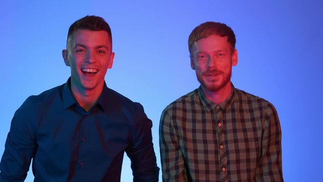 Young attractive homosexual boyfriends look at camera, found something funny, show with finger and laugh. Casually weared brunette and red men in neon color