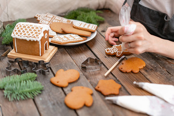 Icing of Christmas bakery. Woman decorating honey gingerbread cookies on wooden brown table. closeup, copy space. Blank biscuit gingerbread house, ready to decorate.