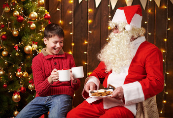 Santa Claus and child boy drinking tea, eating cookies, talking and sitting indoor near decorated xmas tree with lights - Merry Christmas and Happy Holidays!