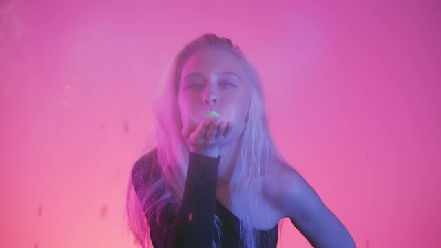 Vivid neon style light. Sexy young woman with with creative make up art face blowing confetti glitter or pieces of gold foil on the camera, it shines brightly in the light