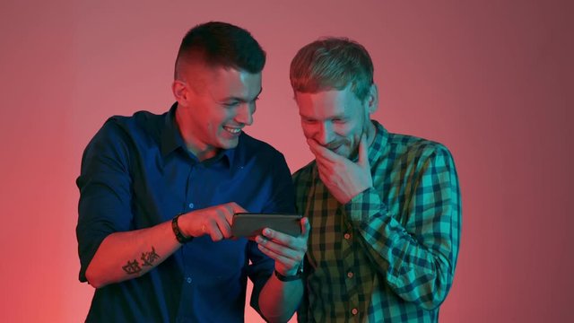 Young attractive homosexual boyfriends watch funny videos and photos on their smartphone. Brunette and red man in neon color light in studio isolated