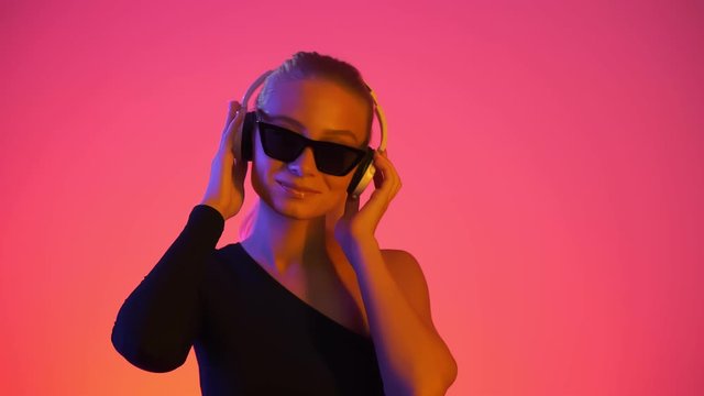 Young blonde woman in sunglasses listens music in headphones at studio. Portrait of happy girl enjoys favorite songs in headphones in vivid neon light