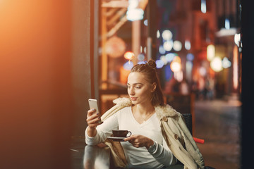 girl drinking coffee and using her phone outside a cafe during the evening