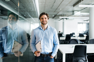 Portrait of young businessman standing in an office, looking at camera. - obrazy, fototapety, plakaty