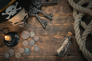 Pirate letter parchment in a bottle, human skull, old coins, moorings, keys and burning candle on a brown wooden table background.