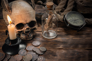 Pirate letter parchment in a bottle, human skull, old coins, compass and burning candle on a brown wooden table background.