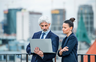 A businessman and businesswoman with laptop standing on a terrace, working.