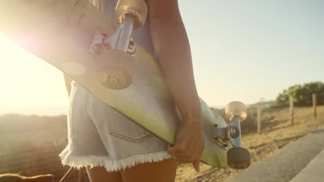 Close up shot of girl holding skateboard walking at sunset with lens flare