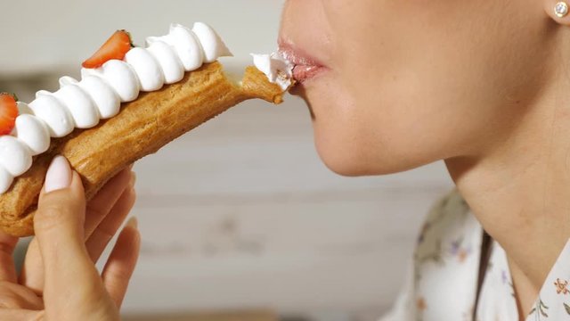 pretty young woman eats beautiful tasty eclair with cream and strawberry slices on blurred background extreme close view