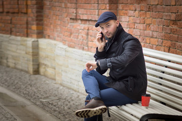 A young man sitiing on a bench and making the call on his phone