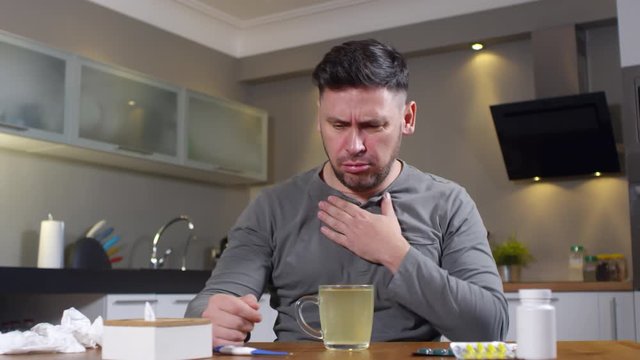 Medium shot of man sitting at kitchen table and having coughing fit, then talking to doctor on video call and complaining about symptoms