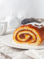 A piece of homemade sponge cake with apple jam. On a white plate. In the background is a white teapot and a mug. Close-up.